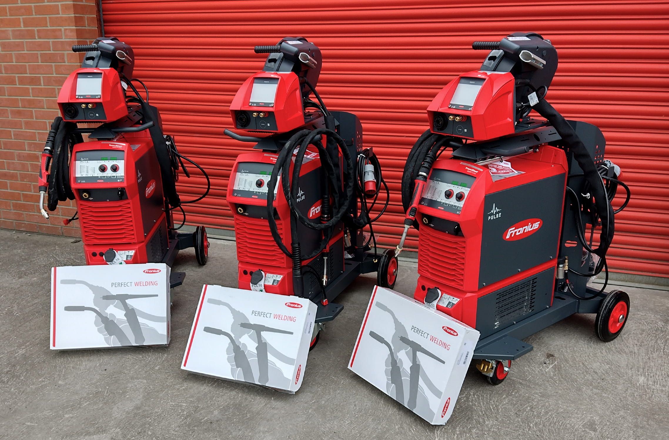 Three watercooled Fronius Transsteel 4000c Pulse machines set up outside a red shutter and awaiting delivery